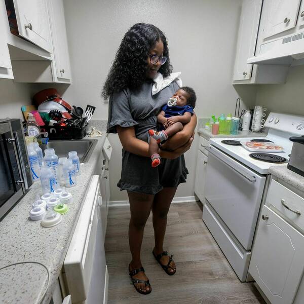 Young woman standing in the middle of a kitchen while holding her infant child with infant bottles and food appliances around.